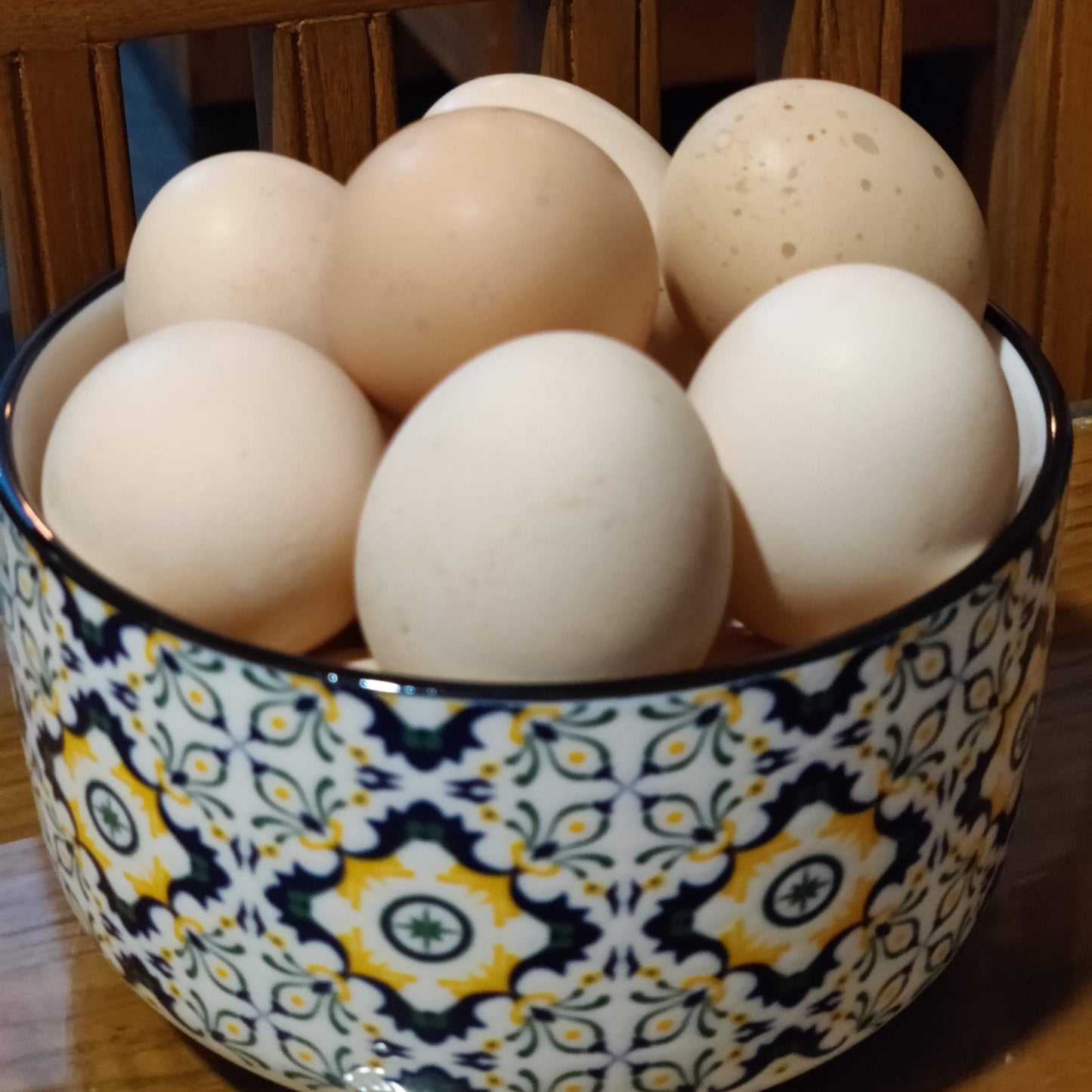 A group of light-colored eggs, including some speckled brown and creamy white eggs, arranged inside a decorative ceramic bowl with a blue, yellow, and white floral pattern, set on a wooden table in front of a wooden chair.